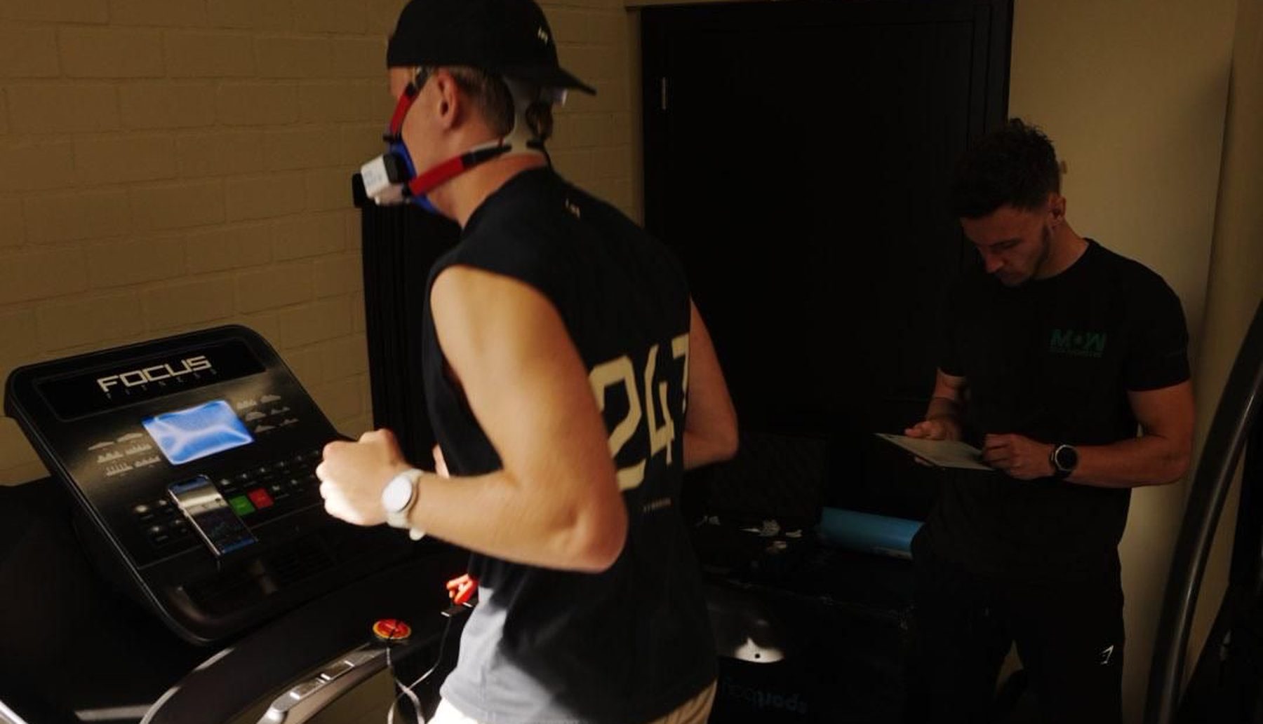 Atleet loopt met ademmasker op de loopband tijdens een VO2max-test, coach van Momentom noteert de meetwaarden
